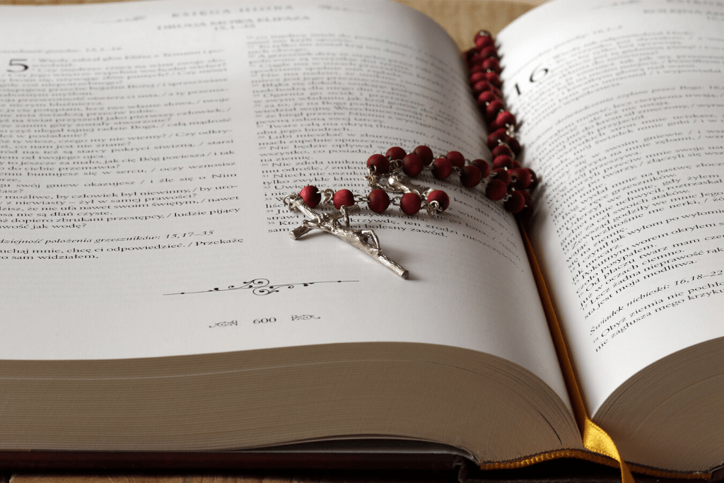 A metal rosary with red beads sits on top of an opened bible