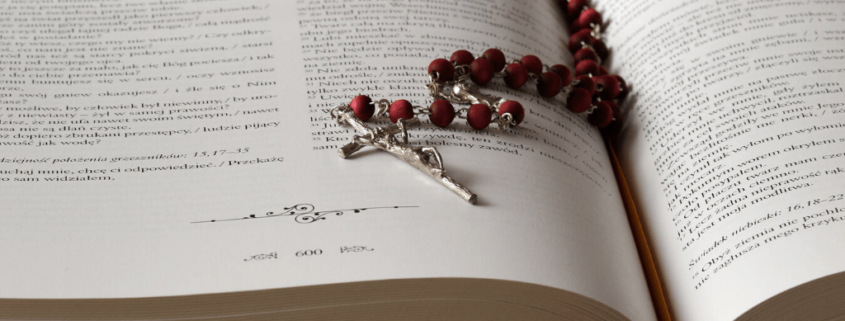 A metal rosary with red beads sits on top of an opened bible