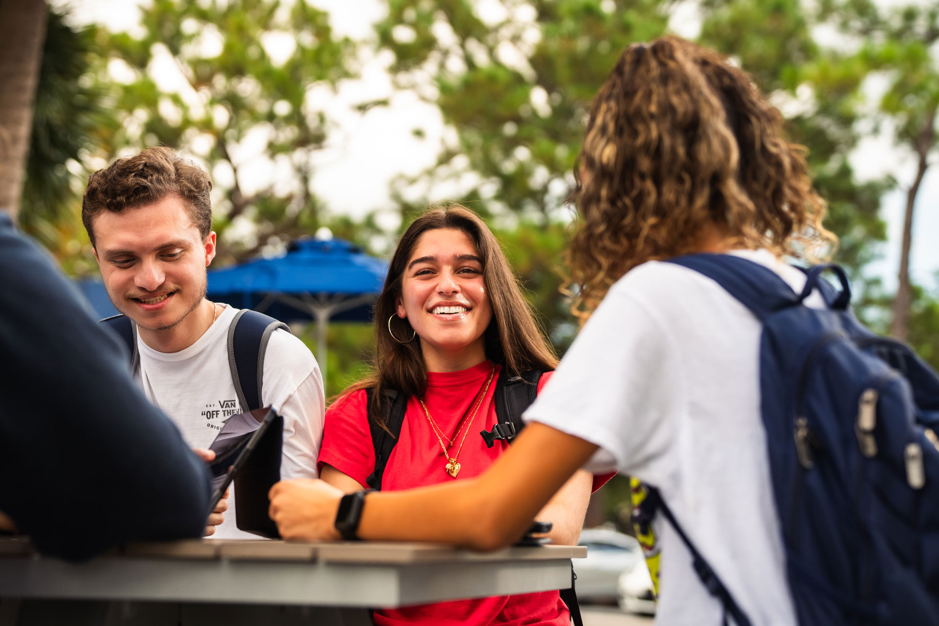 A group of three students is gathered outdoors on a university campus. They are casually interacting around a table. The central focus is on a smiling young woman with long brown hair, wearing a red shirt and a backpack, who is looking directly at the camera. To her left, a young man in a white shirt and a backpack is glancing downward, engaged with something on the table. The third person, whose face is not fully visible, is on the right, also participating in the conversation. The background features trees and a blue umbrella, contributing to a relaxed and friendly atmosphere.