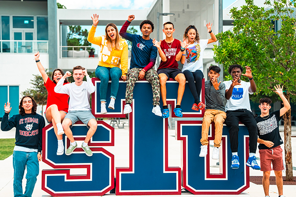 Ten energetic students sit and stand atop the large red, white, and blue "STU" letters on campus. They cheer, wave, and pose playfully, showcasing diverse school pride in front of a modern university building and tree-lined walkway.
