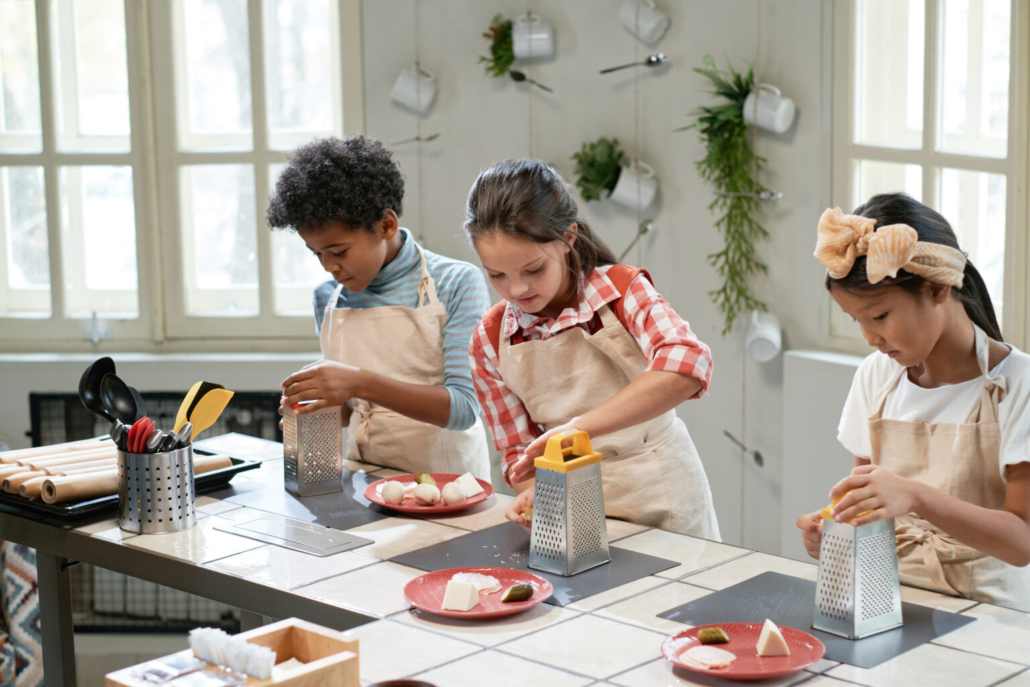Group of children in aprons rubbing cheese on the plates while standing at the table, they cooking pizza during lesson